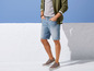 Mann in grüner Jacke, blauen Jeans-Shorts und grauen Sneakern.