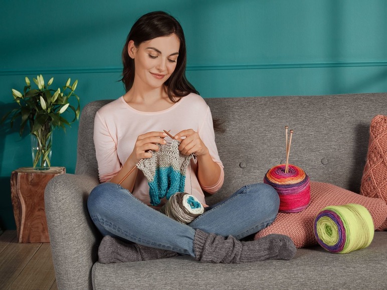 Eine Frau strickt mit bunten Garnen auf einem grauen Sofa.