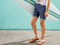 Ein Junge trägt Jeans-Shorts und Flip-Flops und hält ein Surfbrett.