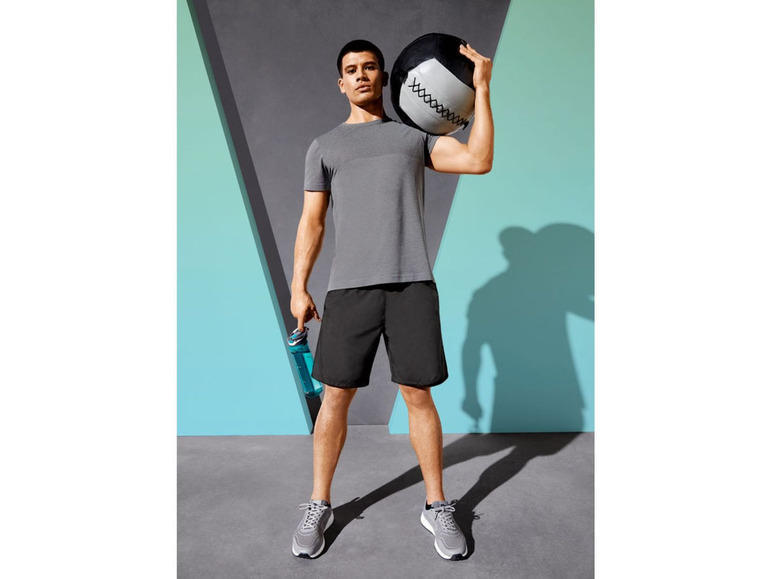 Mann mit grauem Shirt, schwarzen Shorts, Sportschuhen und einem Medizinball.