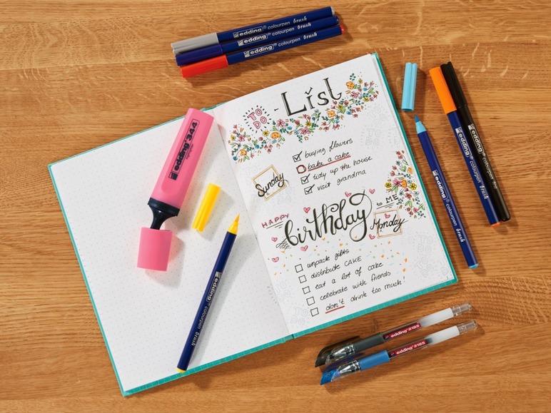 Ein rosa Markierstift und blaue Stifte von Edding auf einem geöffneten Notizbuch.
