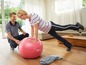 Eine Frau trainiert mit einem pinken Fitnessball, angeleitet von einem Trainer.