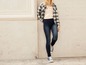 Eine Frau trägt dunkelblaue Skinny Jeans, ein weiß-schwarzes kariertes Hemd und blaue Sneakers.