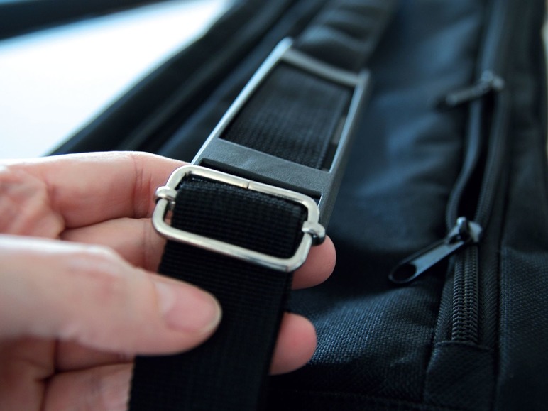 Eine Hand hält den Schultergurt einer schwarzen Tasche mit Reißverschluss fest.
