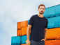 Mann in einem dunkelblauen T-Shirt vor einem Containerterminal.
