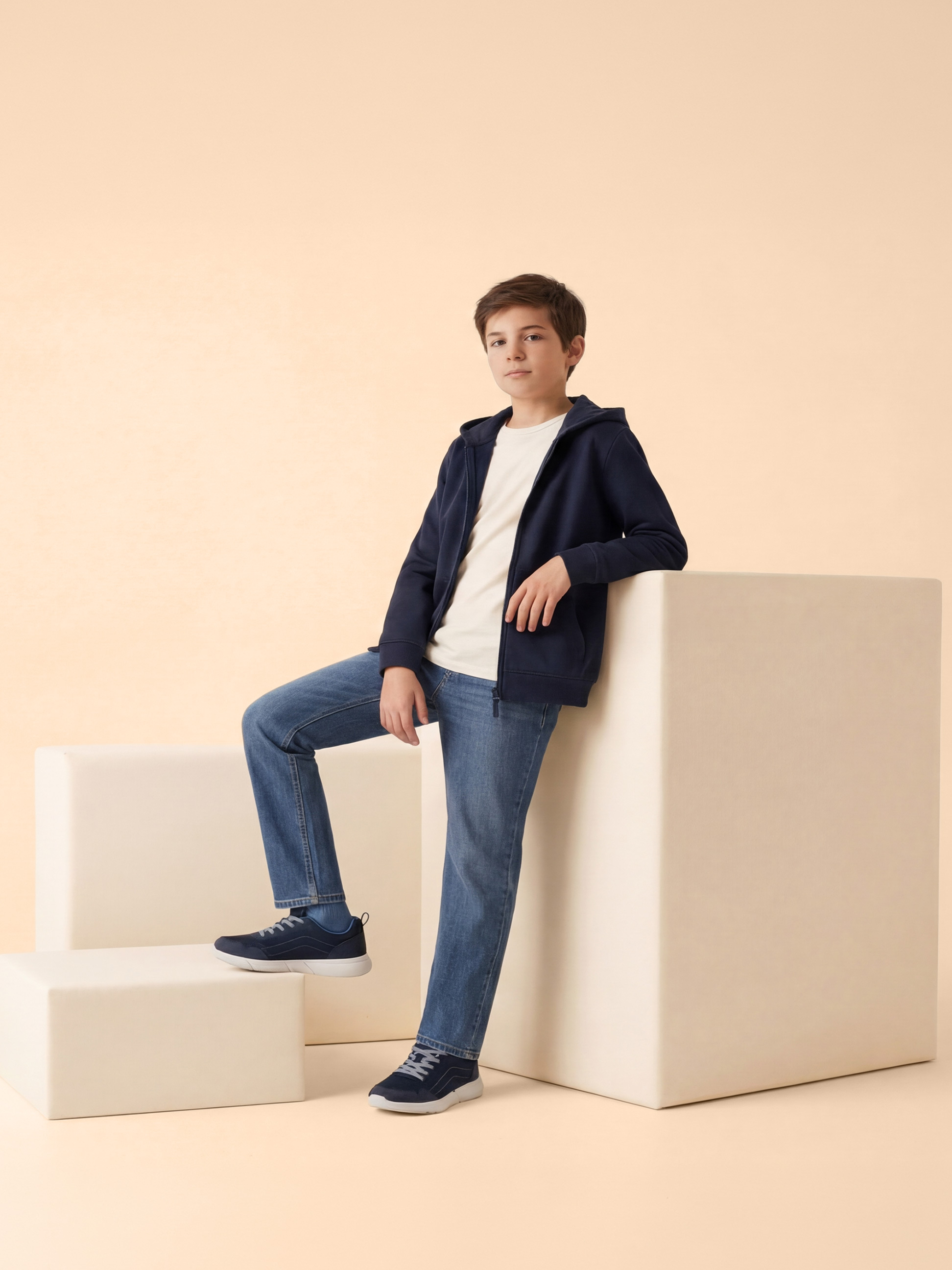 Junge in blauem Kapuzenpullover, weißem T-Shirt und blauen Jeans, lehnt an weißen Blöcken.