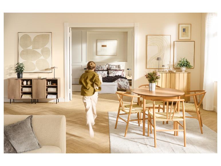 Wohnzimmer mit Essbereich und Schlafzimmer im Hintergrund, Holzmöbel, Bilder, Pflanzen, Teppich.