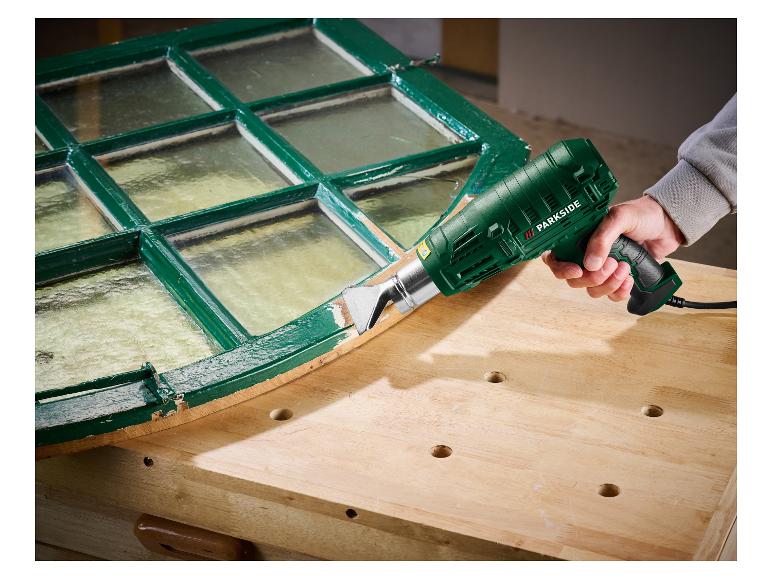 Eine Person verwendet eine Parkside Heißluftpistole, um Farbe von einem grünen Fensterrahmen zu entfernen.