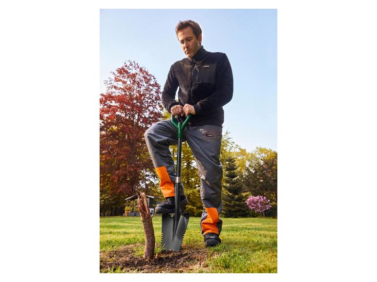 Mann in schwarzer Fleecejacke und orange-grauer Arbeitshose mit Spaten im Garten.