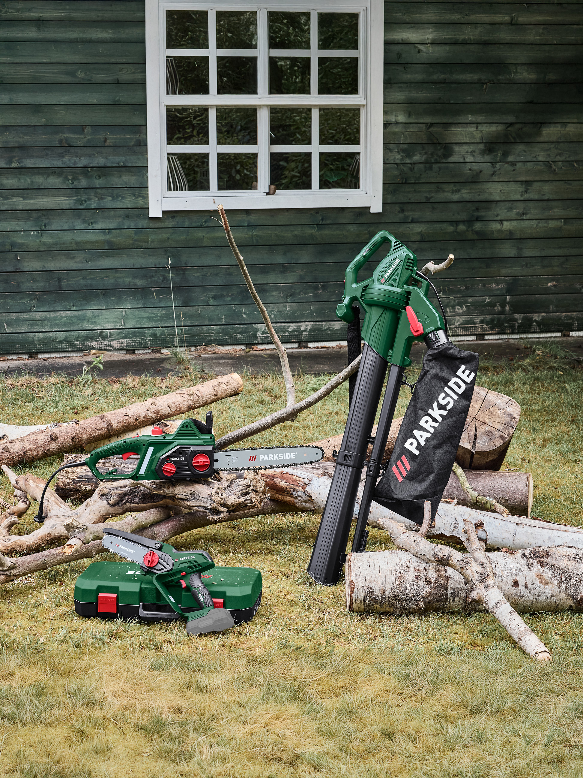 Laubbläser, Kettensäge und Mini-Kettensäge auf Gras neben einem Holzschuppen.