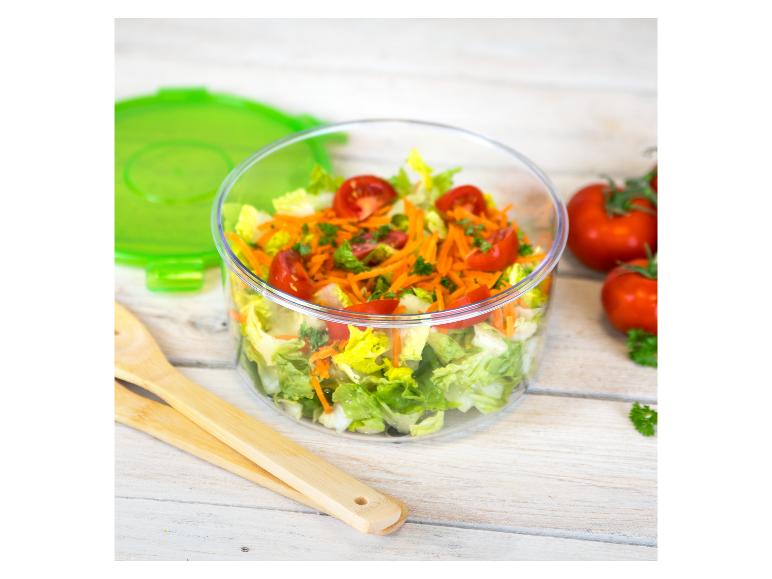 Eine klare Schüssel mit Salat, Tomaten und Karotten, mit grünem Deckel und Holzbesteck auf einem hellen Holztisch.