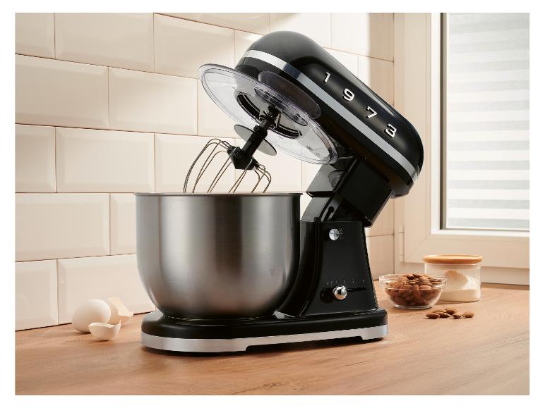 Schwarze Küchenmaschine mit Edelstahlschüssel, Eiern und Mandeln auf einer Holztheke.