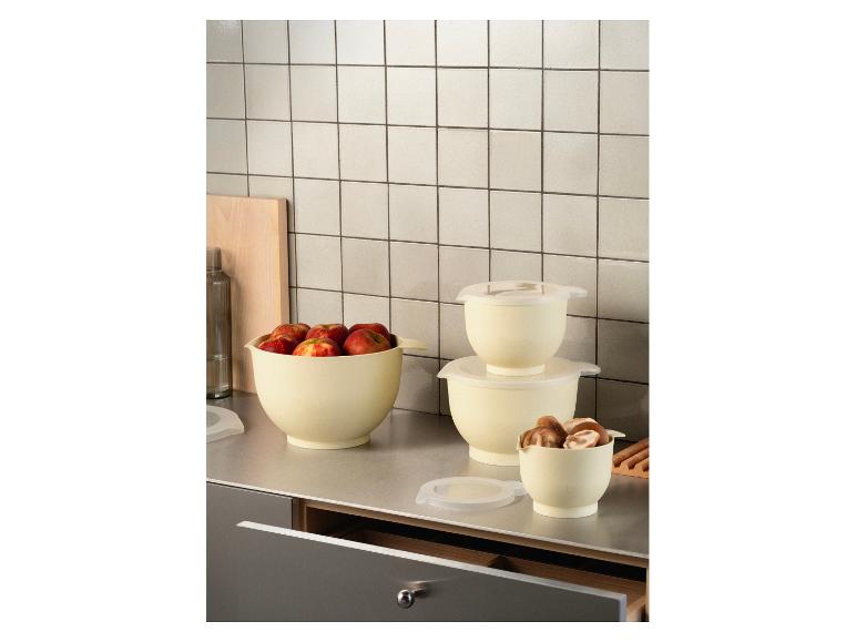 Cremefarbene Rührschüsseln mit Deckeln, eine mit Äpfeln, eine andere mit Pilzen, auf einer Küchenarbeitsplatte.