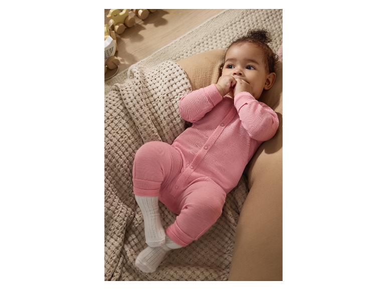 Baby in einem rosa Strampler liegt auf einer Strickdecke, mit Holzspielzeug im Hintergrund.