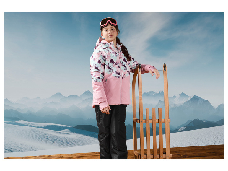 Ein Mädchen in einer rosafarbenen Skijacke und einer schwarzen Skihose steht an einem Schlitten in einer Winterlandschaft.