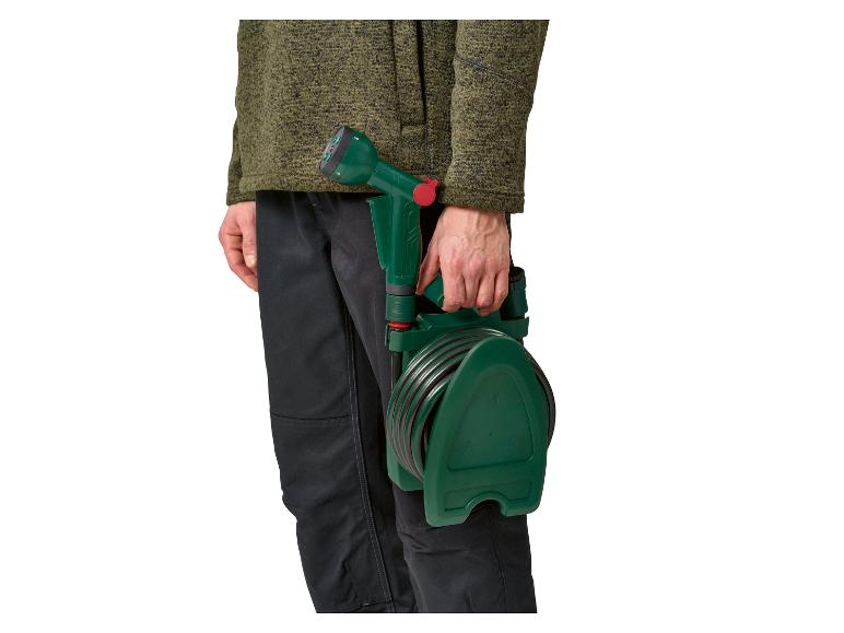 Mann in grüner Jacke und dunkler Hose hält einen Gartenschlauch mit Aufroller.