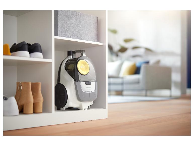 Staubsauger, Schuhe und Aufbewahrungsbox in einem weißen Regal, mit unscharfem Wohnzimmer im Hintergrund.