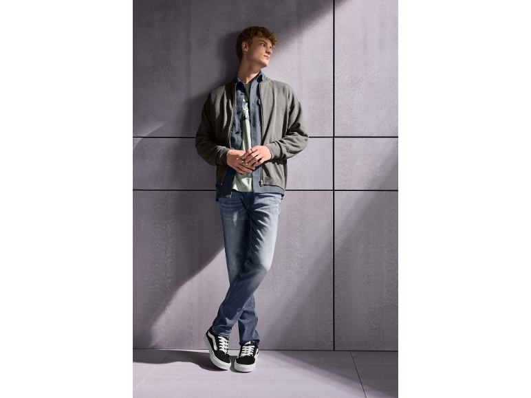 Junger Mann in grauer Jacke, Jeans und Turnschuhen, an eine Wand gelehnt.