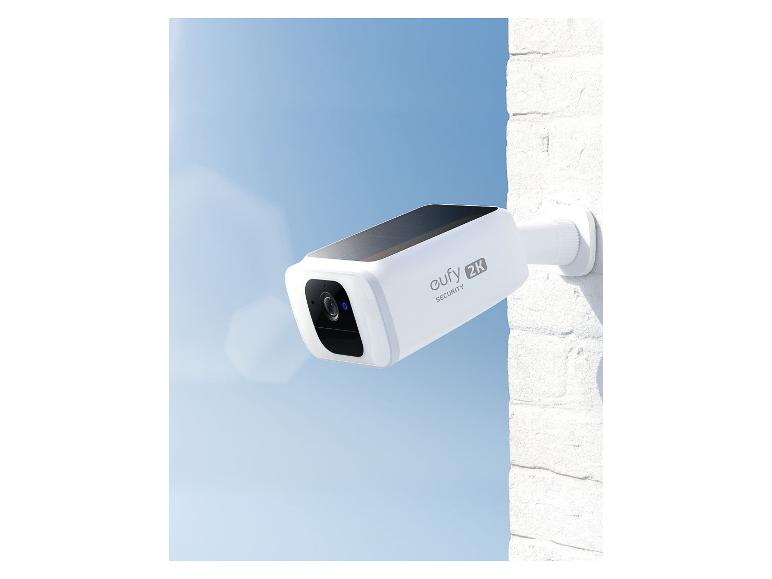 Weiße Überwachungskamera mit Solarpanel an einer Ziegelwand unter blauem Himmel.