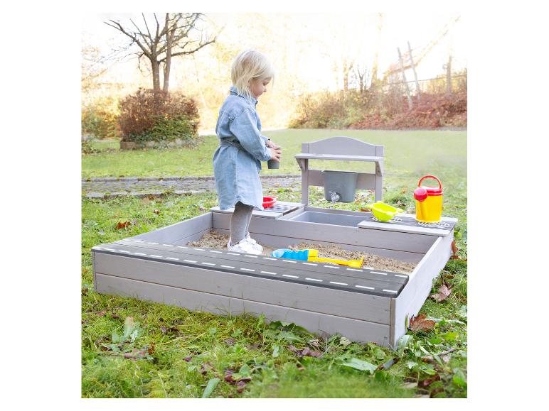 Heller Holzsandkasten mit Spielküche und Spielzeug, spielendes Mädchen im Freien.