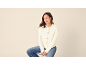 Frau in cremefarbener Strickjacke und Jeans sitzt auf einem Hocker, blickt nach rechts.