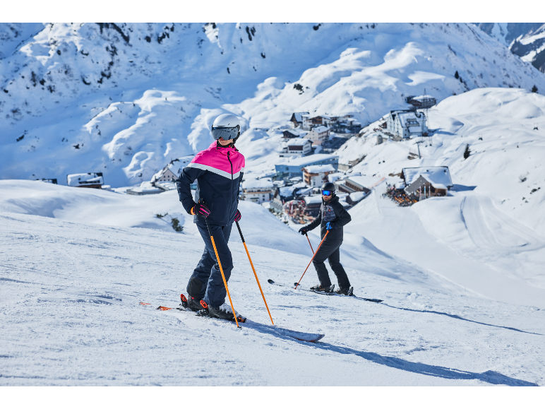 Zwei Skifahrer in Skikleidung und Helmen auf einem verschneiten Berghang mit Dorf im Hintergrund.