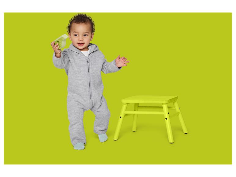 Baby im grauen Strampler mit grünem Spielzeug und gelbem Hocker auf grünem Hintergrund.