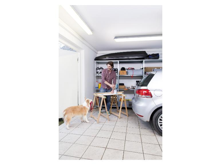 Mann streicht ein Holzbrett in einer Garage, mit Hund und Auto daneben.