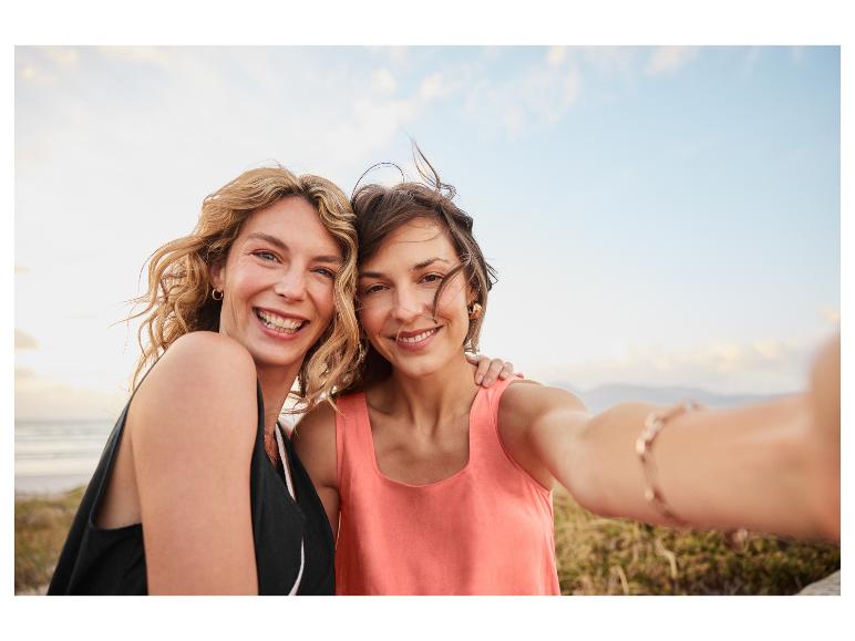 Zwei lächelnde Frauen in Sommerkleidern am Meer.