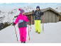 Zwei Kinder in Winterkleidung mit Skistöcken in einer verschneiten Berglandschaft mit Holzhütte.