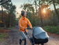 Eine Frau in einer orangenen Jacke joggt mit einem Kinderwagen durch einen Wald.