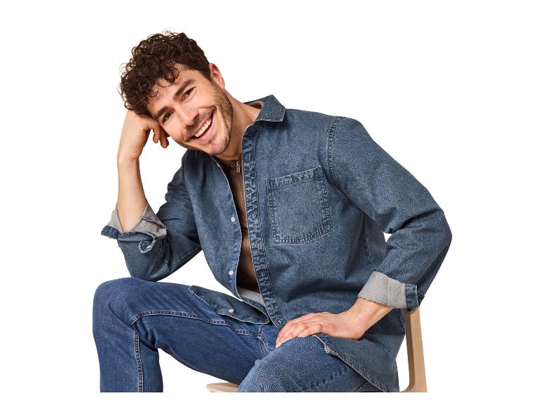 Lächelnder Mann in Jeanshemd und Jeans, auf einem Stuhl sitzend.