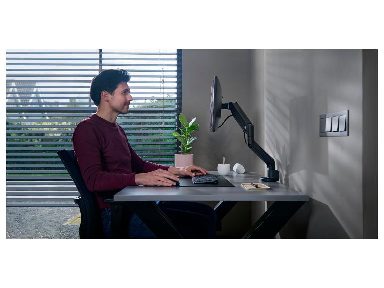 Mann arbeitet am Schreibtisch mit Monitor, Tastatur und Maus, neben Fenster mit Jalousien.