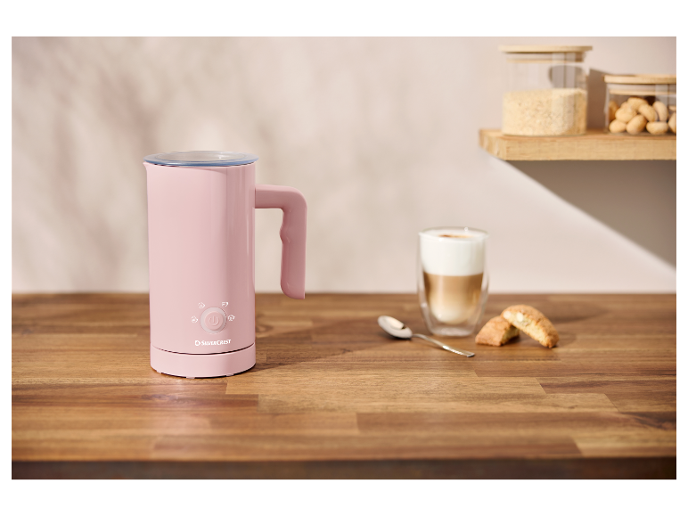 Ein rosa Milchaufschäumer von SilverCrest auf einem Holztisch, neben einem Cappuccino und Keksen.
