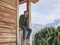 Mann in grünem Pullover und blauer Jeans sitzt auf einem Holzbalkon mit Blick auf die Berge.