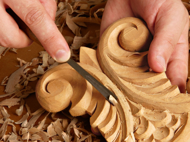 Eine Hand bearbeitet ein Holzornament mit einem Meißel.