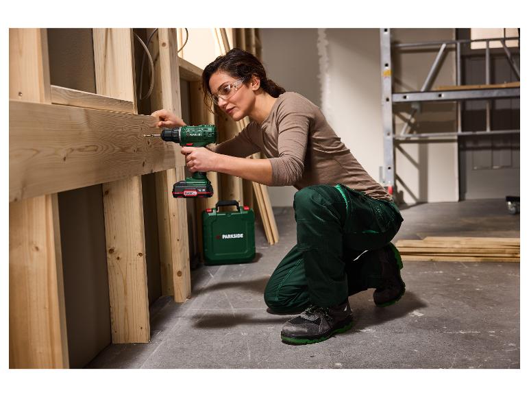 Frau in Schutzbrille und Arbeitskleidung benutzt Akkuschrauber an Holzrahmen.