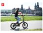 Frau mit Helm fährt ein Klapp-E-Bike auf einem Radweg mit Fluss und Gebäuden im Hintergrund.