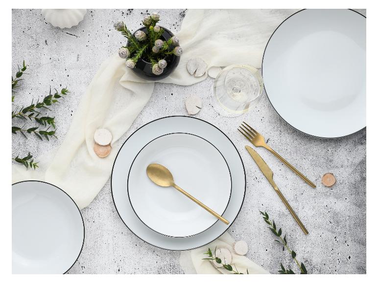 Gedeckter Tisch mit weißen Tellern, goldenem Besteck, Glas und grünen Zweigen auf grauem Hintergrund.