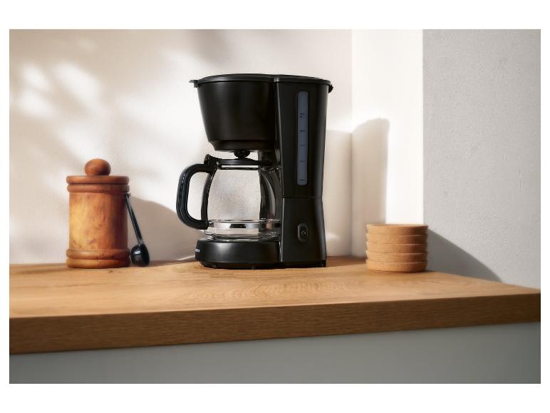 Schwarze Filterkaffeemaschine mit Glaskanne, Holzgefäß und Korkuntersetzern auf einer Theke.