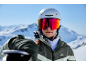 Skifahrerin mit Helm und Skibrille, Skier auf der Schulter, vor Bergpanorama