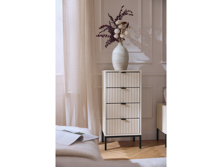 Helle Kommode mit vier geriffelten Schubladen und einer Blumenvase darauf.