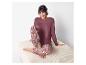 Frau in bordeauxrotem Pyjama und gemusterter Hose sitzt auf einem hellen Hocker.