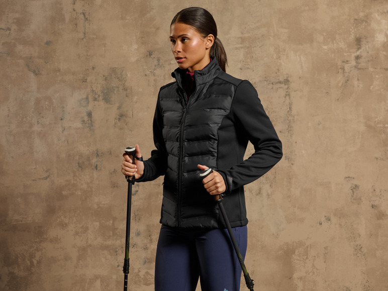 Eine Frau in einer schwarzen Jacke und blauen Hosen hält Wanderstöcke.