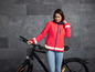 Eine Frau in einer leuchtend rosa Jacke mit reflektierenden Streifen steht neben einem schwarzen Fahrrad.
