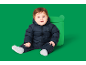 Baby in einem dunkelblauen wattierten Schneeanzug mit Bären-Emblem, sitzend neben einer grünen Spielzeugkiste.
