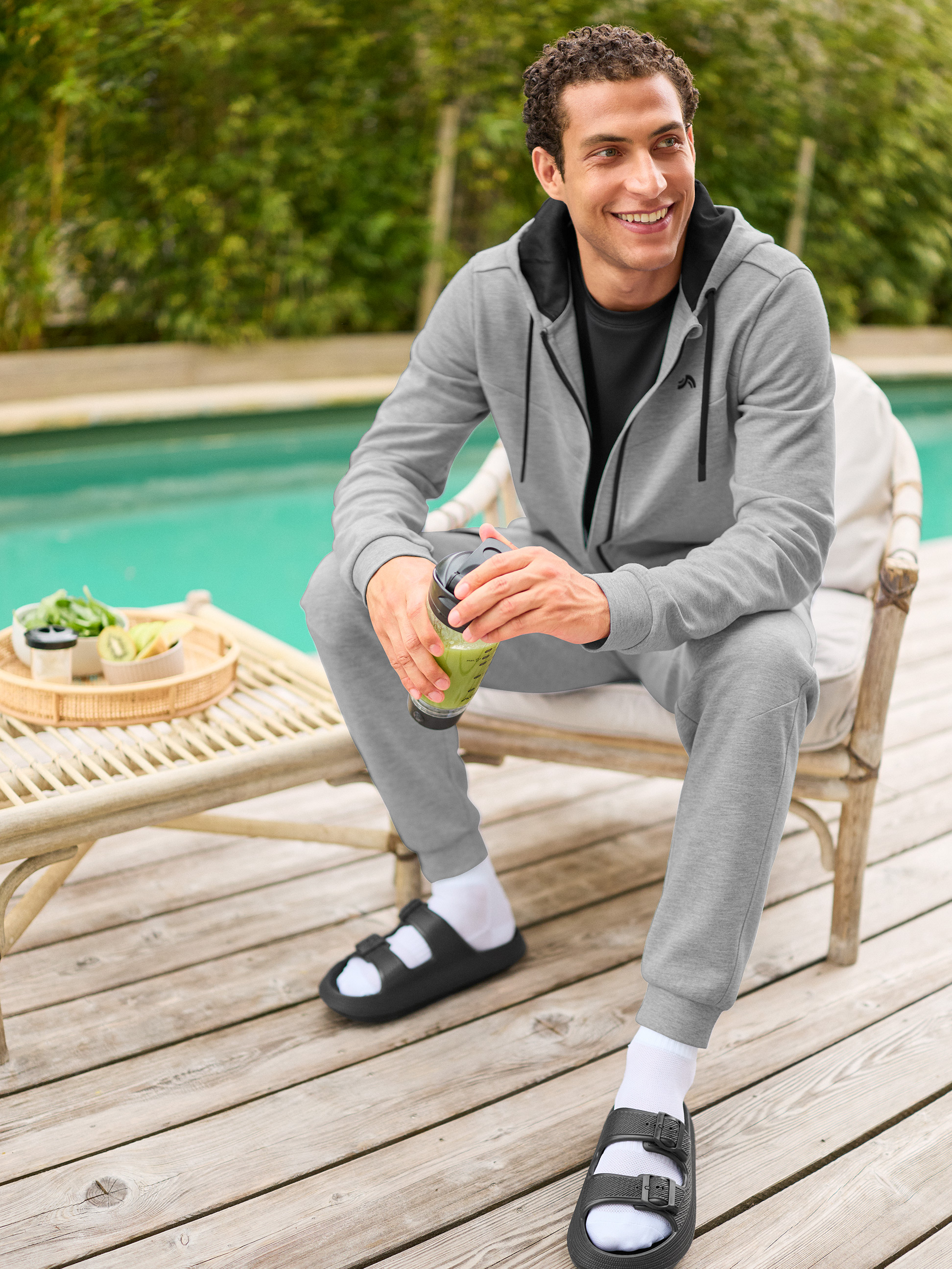 Mann in grauem Trainingsanzug und schwarzen Sandalen mit weißen Socken, hält einen grünen Smoothie am Pool.