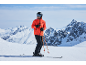 Mann in orangefarbener Skijacke, schwarzer Skihose, Helm und Skibrille auf einem verschneiten Berggipfel.