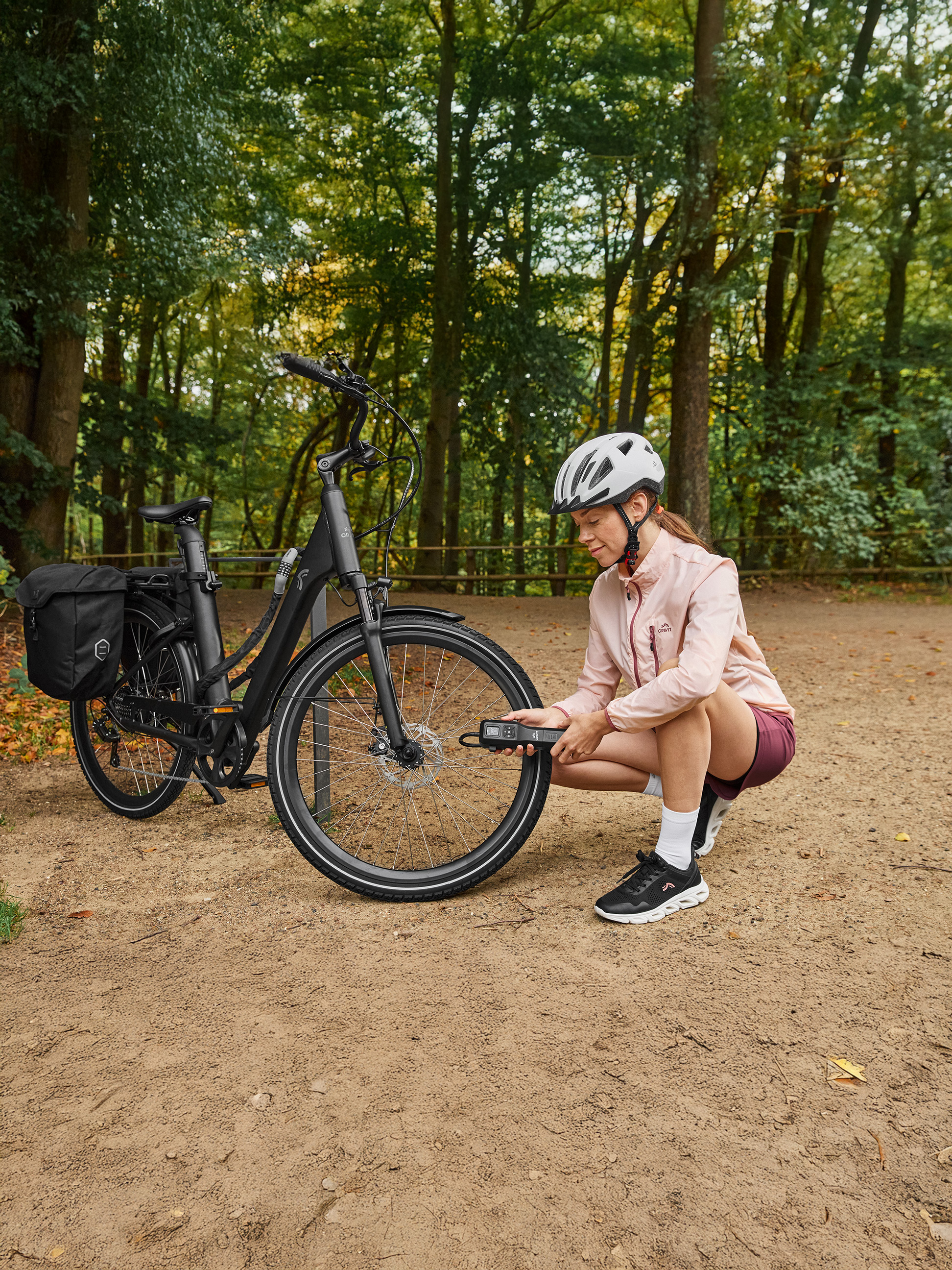 Frau in Crivit Sportkleidung prüft E-Bike-Reifendruck mit einer tragbaren Pumpe im Wald.