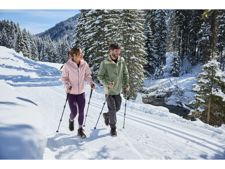 Zwei Personen mit Wanderstöcken gehen auf einem verschneiten Weg zwischen Tannen.